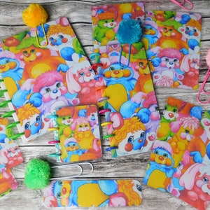 May include: A collection of colorful notebooks and notepads with a repeating pattern of cartoon characters. The notebooks have spiral bindings in various colors, and some are accented with pom-pom paperclips. The characters are in shades of blue, pink, yellow, and green.