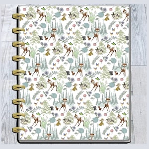 May include: A planner cover with a whimsical forest pattern featuring deer, rabbits, butterflies, and trees in shades of brown, green, and blue on a white background. The planner has gold-colored disc binding.