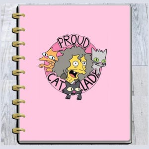 May include: A pink planner with gold-coloured binding rings. The cover features a cartoon image of a woman with grey hair and three cats, with the words "PROUD CAT LADY" in a pink circle.