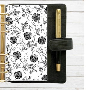 Puede incluir: Agenda de cuero negro con un bolígrafo dorado y una inserción con estampado floral. La inserción presenta un diseño floral en blanco y negro con grandes flores tipo amapola y hojas. La agenda tiene una encuadernación dorada.