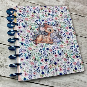 May include: A floral patterned notebook with blue binding rings. The cover features a colorful design of flowers and foliage, with an illustration of Bambi and Thumper. The notebook is open, revealing the interior pages.