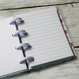 May include: Open notebook with lined paper and a red top line. The pages are held together by four white plastic disc-bound rings. The cover has a pink pattern with the letter "Y".