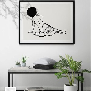 Puede incluir: Impresi&oacute;n en blanco y negro de arte de l&iacute;nea de una mujer con afro tumbada sobre un fondo blanco. La impresi&oacute;n est&aacute; enmarcada en un marco negro y colgada en una pared blanca.