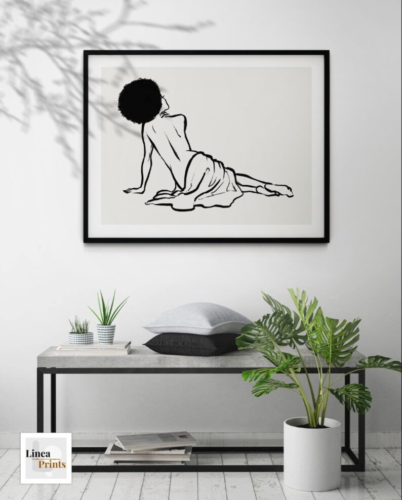 Puede incluir: Impresi&oacute;n en blanco y negro de arte de l&iacute;nea de una mujer con afro tumbada sobre un fondo blanco. La impresi&oacute;n est&aacute; enmarcada en un marco negro y colgada en una pared blanca.