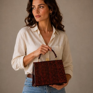 May include: A rectangular handbag with a snakeskin pattern in shades of brown and red. The bag has a gold-coloured clasp and handle attachments. The handle is a matching brown colour. The bag is held by a person wearing a white shirt and blue jeans.