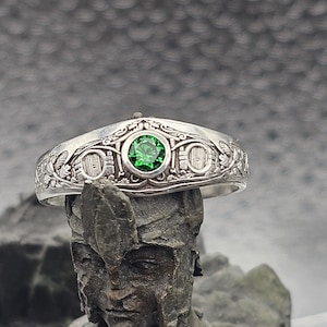 May include: A silver ring with an intricate design featuring a green gemstone. The ring is set on a textured surface.