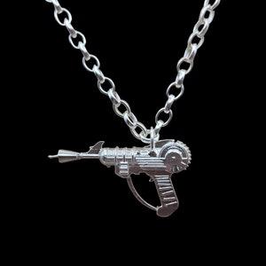 May include: A silver necklace featuring a detailed, futuristic-looking toy gun pendant. The gun has a barrel, a handle, and a circular detail on the side.