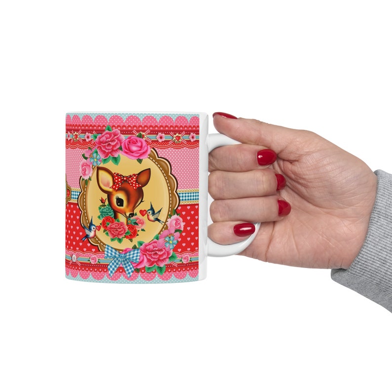 May include: White ceramic mug with a pink, red, and blue floral design featuring a cartoon deer with a red bow. The design includes hearts, birds, and a scalloped border.