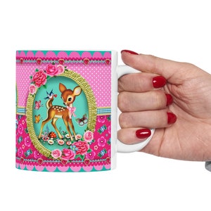 May include: White ceramic mug with a pink, green, and gold design featuring a cartoon deer, flowers, and birds. The mug has a scalloped edge and a heart pattern.