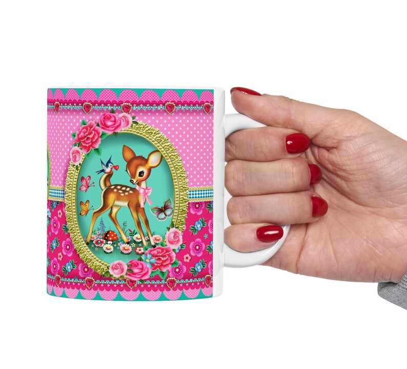 May include: White ceramic mug with a pink, green, and gold design featuring a cartoon deer, flowers, and birds. The mug has a scalloped edge and a heart pattern.