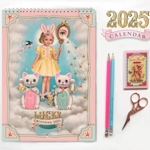May include: A pink and blue calendar with a whimsical illustration of a girl with bunny ears, two white cats, and stars. The calendar is titled "Lucky Calendar 2025".