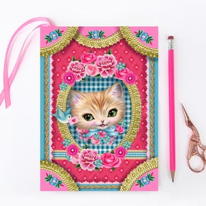 May include: A pink notebook with a decorative frame featuring a kitten illustration, pink ribbon, pink pencil, and rose gold scissors. The frame has gold trim, pink roses, and a blue bird holding a letter. The background is pink and blue.