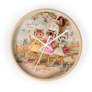 May include: A round wall clock with a light wood frame. The clock face features a vintage illustration of three anthropomorphic cats in dresses, with the text "Greetings from Happy Island". The background includes a tree, flowers, and butterflies.