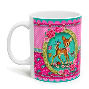 May include: A white ceramic mug with a pink, blue, and green floral design. The design features a deer with a butterfly and birds in a gold frame.