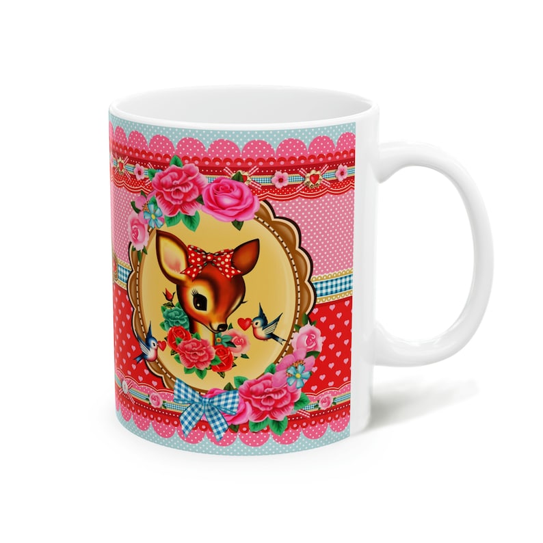 May include: White ceramic mug with a colorful design featuring a cartoon deer with a red bow, surrounded by pink and red flowers and blue birds. The background is a pink and red polka dot pattern with white lace trim.