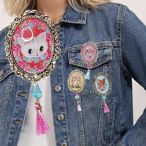 May include: Four oval-shaped brooches with ornate gold-tone metal frames. Each brooch features a vintage-style illustration of a kitten, fawn, or bunny surrounded by flowers. Each piece includes a dangling blue crystal bead and a pink fabric tassel.