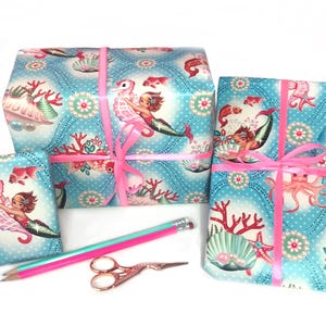 May include: Three gift boxes wrapped in blue and white wrapping paper with a mermaid and seahorse design. The boxes are tied with pink ribbon.