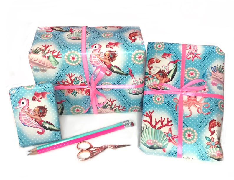 K&ouml;nnte beinhalten: Drei Geschenkboxen, verpackt in blaues und wei&szlig;es Geschenkpapier mit einem Meerjungfrauen- und Seepferdchen-Motiv. Die Boxen sind mit einem rosa Band gebunden.