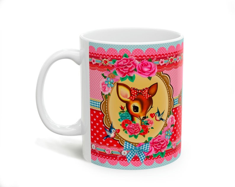 May include: White ceramic mug with a pink, red, and blue floral design featuring a cartoon deer with a bow. The design includes birds and hearts.