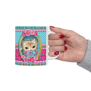 May include: White ceramic mug with a colorful design featuring a kitten in a blue and white checkered frame surrounded by pink and blue flowers. The mug has a gold trim.