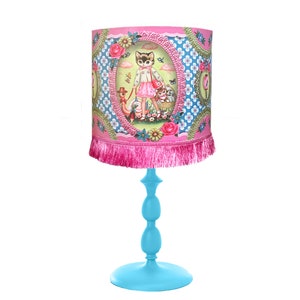 Puede incluir: Una lámpara de mesa azul con una pantalla rosa que presenta un gato y otros animales en una escena de jardín caprichosa. La pantalla tiene un ribete de flecos rosa.