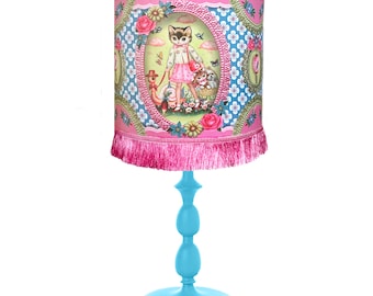 May include: A blue table lamp with a pink lampshade featuring a cat and other animals in a whimsical garden scene. The lampshade has a pink fringe trim.