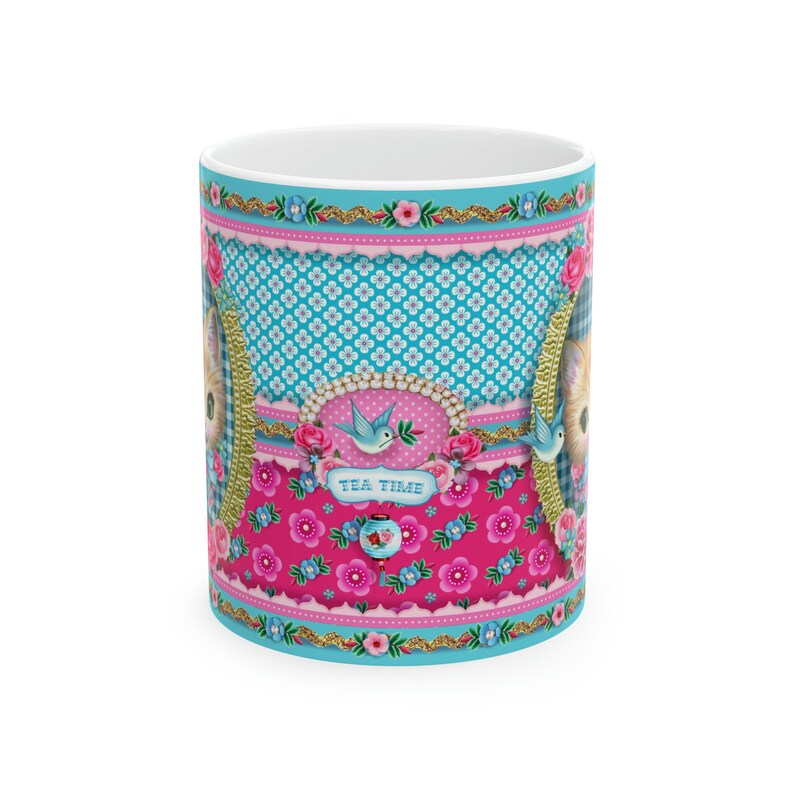 May include: A white ceramic mug with a colorful design featuring a pink and blue floral pattern, a blue bird, and a cat's face. The text "Tea Time" is printed on the mug.