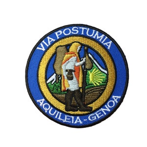 Può includere: Una toppa ricamata blu e oro con il testo "VIA POSTUMIA AQUILEIA - GENOA" e la raffigurazione di un pellegrino che cammina con un bastone e uno zaino.