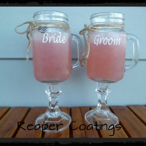 May include: Two clear glass mason jar style goblets filled with pink liquid. The jars have handles, lids, and are adorned with twine and a decorative charm. One jar reads "Bride" and the other reads "Groom". The base of the goblets are clear glass.