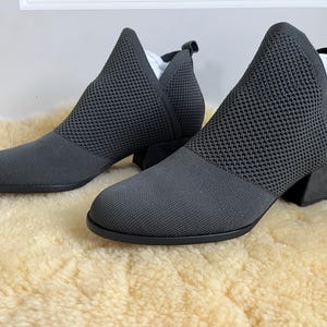 May include: A pair of dark gray ankle boots featuring a textured knit upper, a rounded toe, a V-shaped cutout at the ankle, a pull tab at the heel, and a low block heel. The boots have a solid dark gray base and a matching knit fabric construction.