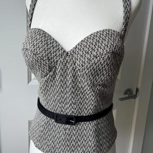 May include: A sleeveless bodice top featuring a black and white herringbone pattern with a sweetheart neckline and thick shoulder straps. The garment is cinched at the waist with a thin black belt that includes a rectangular buckle and metal eyelets.
