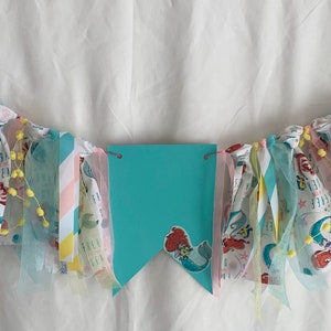 以下が含まれることがあります： 人魚をテーマにしたターコイズと白の布製バナー。バナーの中央には大きなターコイズ色のペナントがあり、その中には小さな人魚の画像があります。バナーは黄色のポンポンとカラフルなリボンで飾られています。
