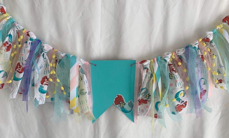 以下が含まれることがあります： 人魚をテーマにしたターコイズと白の布製バナー。バナーの中央には大きなターコイズ色のペナントがあり、その中には小さな人魚の画像があります。バナーは黄色のポンポンとカラフルなリボンで飾られています。