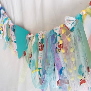 以下が含まれることがあります： 人魚をテーマにしたカラフルな布製のガーランド。ガーランドは、チュール、サテン、コットンなど、さまざまな生地で作られています。ガーランドは、黄色のポンポンと、