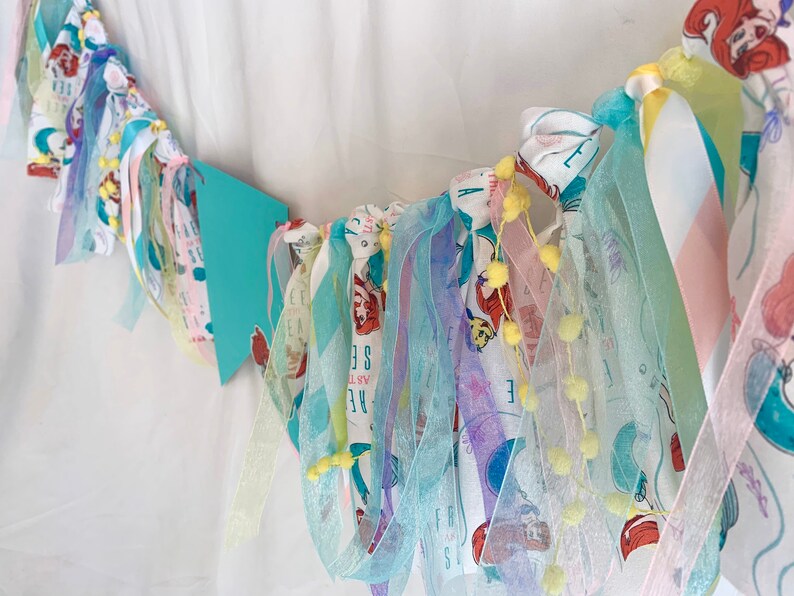 以下が含まれることがあります： 人魚をテーマにしたカラフルな布製のガーランド。ガーランドは、チュール、サテン、コットンなど、さまざまな生地で作られています。ガーランドは、黄色のポンポンと、