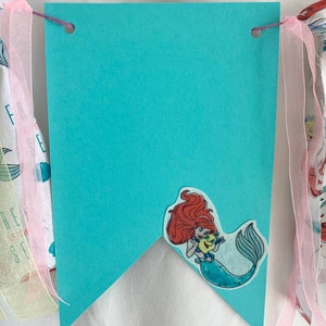 以下が含まれることがあります： アリエルがセバスチャンを手に持っている、ターコイズブルーの旗。旗の上部には、紐を通すための穴が2つ開いています。