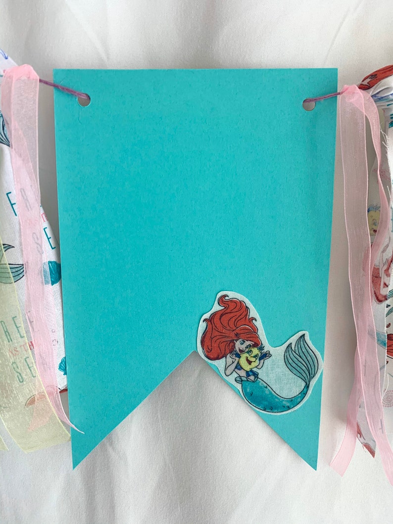 以下が含まれることがあります： アリエルがセバスチャンを手に持っている、ターコイズブルーの旗。旗の上部には、紐を通すための穴が2つ開いています。