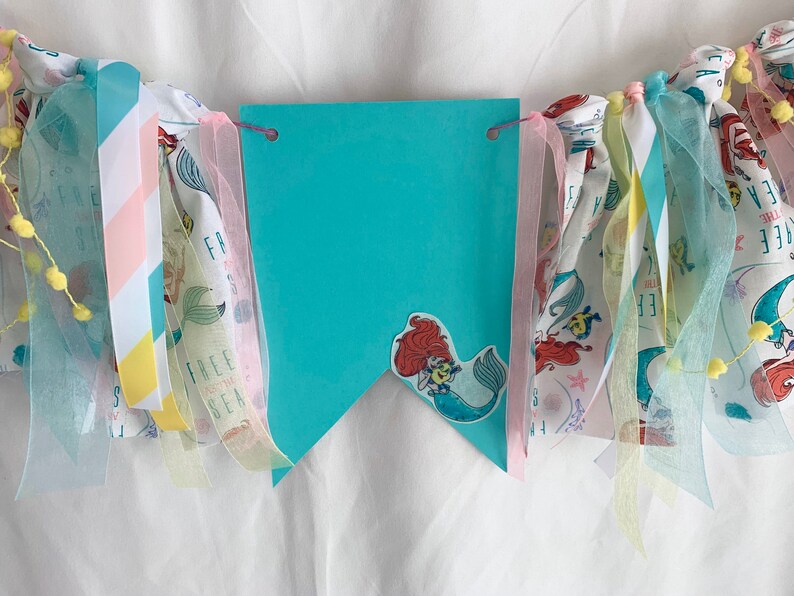 以下が含まれることがあります： ターコイズブルーの旗に、人魚のステッカーと「Free to be the Sea」の文字が書かれています。旗はピンク、イエロー、ブルーのリボンと布の切れ端で飾られています。