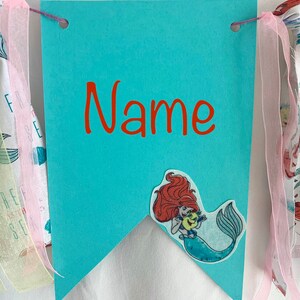 以下が含まれることがあります： ターコイズブルーの旗に赤い文字で「Name」と書かれています。リトルマーメイドのArielの布製アップリケが旗に付いています。