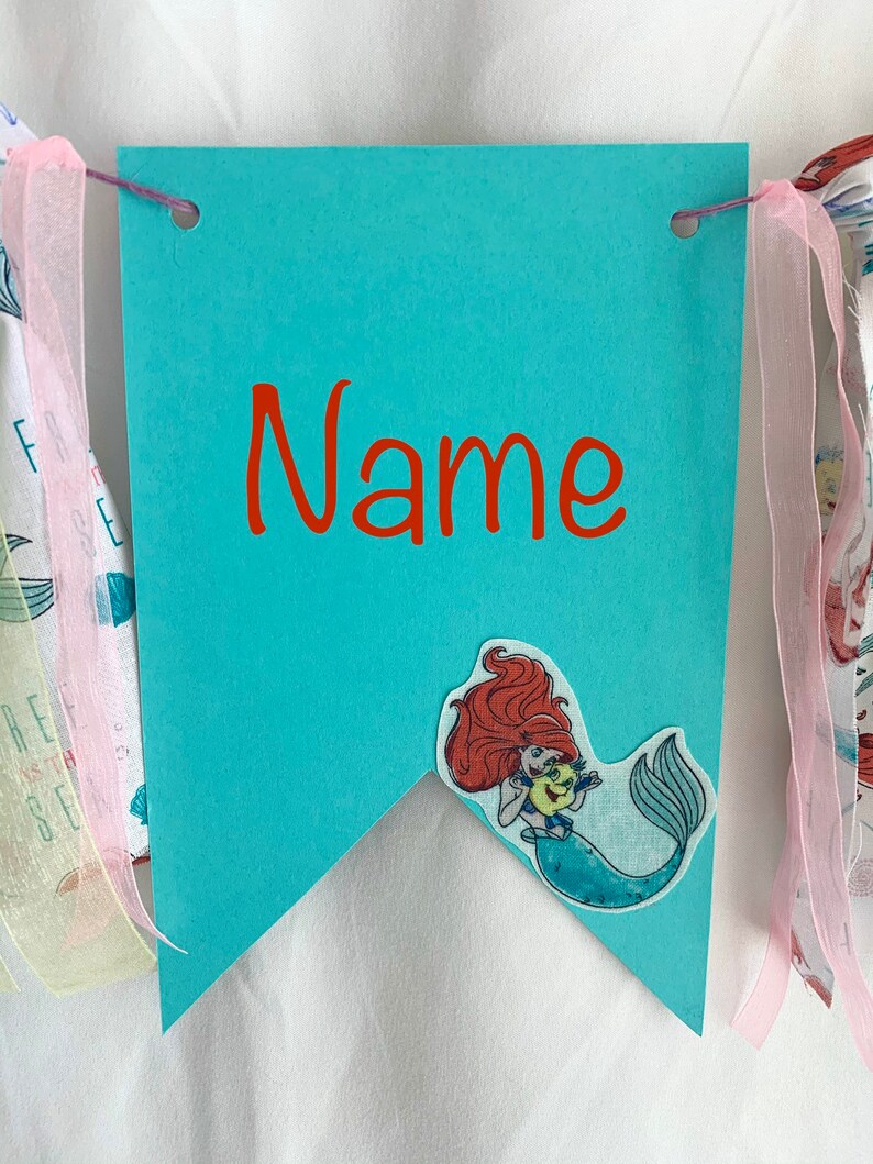 以下が含まれることがあります： ターコイズブルーの旗に赤い文字で「Name」と書かれています。リトルマーメイドのArielの布製アップリケが旗に付いています。