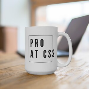 May include: White ceramic mug with the text "PRO AT CSS" in a black, bold font, centered within a black square. The mug has a large handle and is set on a wooden surface, with a laptop visible in the background.