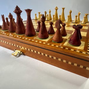 May include: A wooden chess set with a dark brown, inlaid wooden board and matching pieces. The chess pieces are a mix of dark brown and light tan. The set is in a closed wooden box with a gold-coloured clasp.