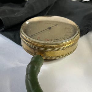 Puede incluir: Un manómetro antiguo de latón con una esfera de cristal abovedada transparente. El medidor tiene un diseño circular con una escala marcada de 0 a 300. Un tubo flexible verde se extiende desde el lateral. Un paño negro está en el fondo.