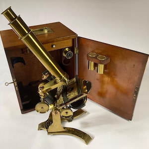 May include: A brass microscope with a polished finish, featuring an adjustable eyepiece, a stage with clips, and multiple focus knobs. It is positioned in front of an open wooden storage case that contains additional lens components and accessories.
