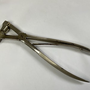 Pode incluir: Uma ferramenta vintage de metal prateado com um gancho curvo e cabos longos e pontiagudos. A ferramenta tem um design articulado e um mecanismo de travamento. O design geral sugere que é um instrumento especializado, possivelmente para um ofício ou comércio específico.