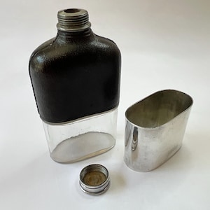 Puede incluir: Un frasco de vidrio transparente con una envoltura de cuero negro y una tapa de metal plateado. También hay una taza de metal plateado separada y una tapa plateada pequeña. El frasco está diseñado para contener líquidos.