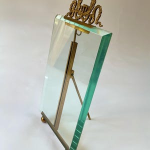 May include: A clear glass picture frame with a brass-colored metal stand and decorative top. The frame has a rectangular shape and is supported by a hinged stand. The decorative top features an ornate design.