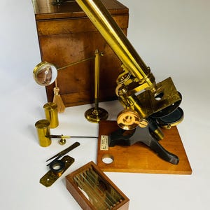 Puede incluir: Un microscopio antiguo de latón con base negra montado sobre una plataforma de madera, acompañado de un estuche de madera, un soporte de latón con lente circular, dos botes de latón, unas pinzas, un portaobjetos de metal y una caja de madera.