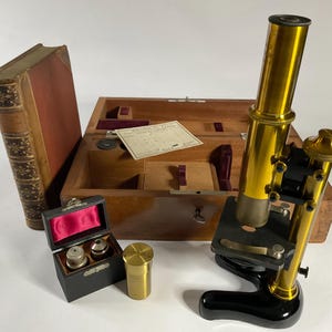 Puede incluir: Un microscopio antiguo con cuerpo de latón y base negra, junto a una caja de madera con accesorios y un libro encuadernado en cuero. El microscopio está inclinado, mostrando su lente y perillas de ajuste. La caja está abierta, revelando compartimentos y un documento.