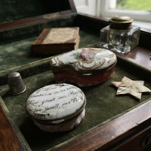 May include: A collection of antique items including two porcelain trinket boxes, one with a floral motif and one with cursive text, a silver thimble, a small leather-bound book, a glass inkwell with a brass lid, and a folded paper star.
